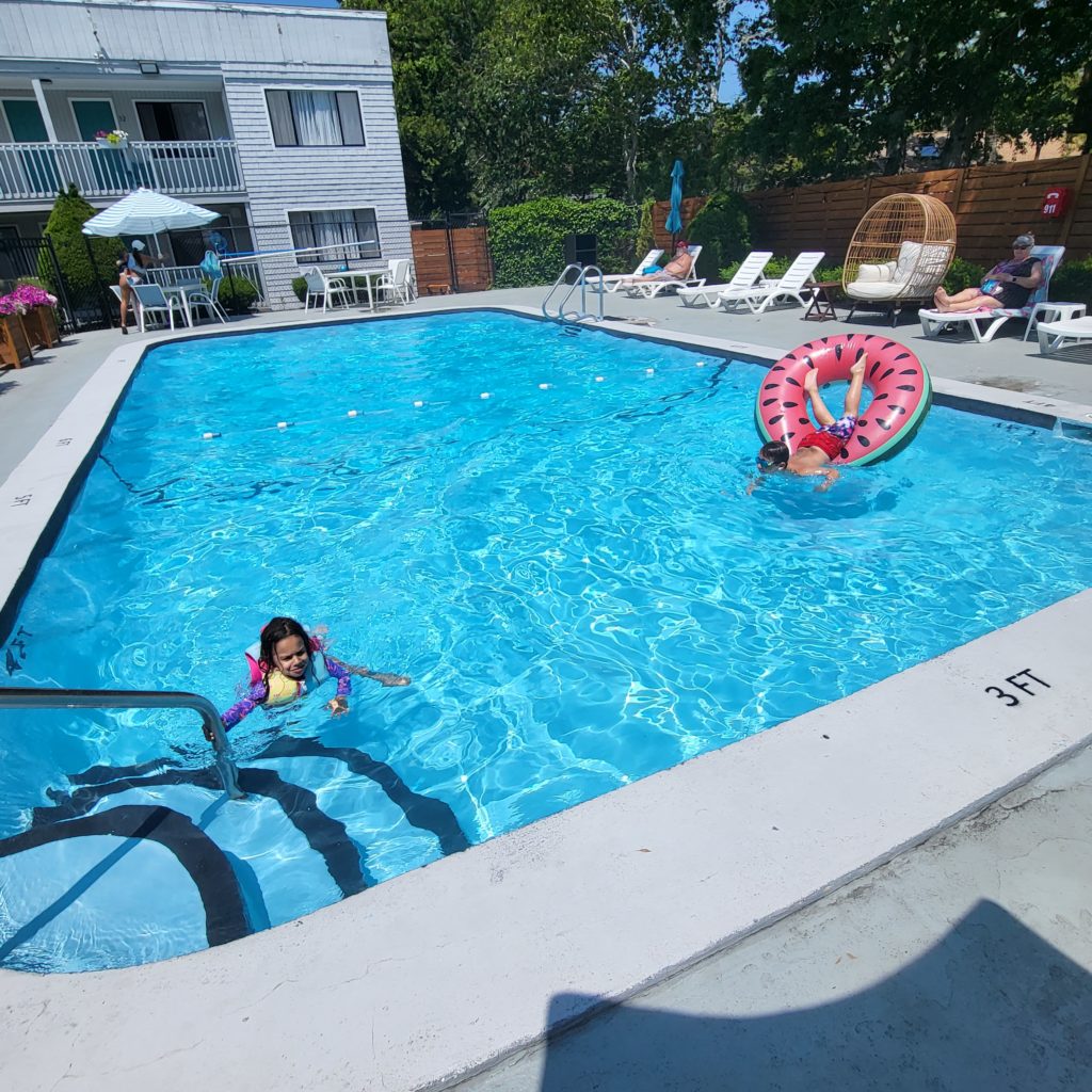 A hotel pool with blue shimmering water, and a rope with oval plastic buoys separating the deep end from the shallow end. The shallow end is in the foreground, marked as 3 FT. A three-year-old in a yellow and blue life jacket, and purple long sleeve bathing suit, is in the pool and holding on to a handrail by the pool stairs. A 6-year-old is across the pool horizontally, still in the shallow end, slipping headfirst from an inner tube decorated like a watermelon. The 6-year-old is wearing red and blue swim trunks and goggles.