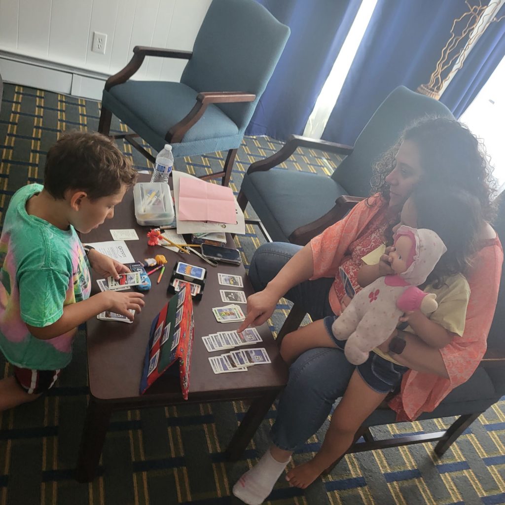 An adult and child playing a card game on a coffee table in a hotel room. The child is kneeling and looking at a card holder with 6 cards in it. The adult is pointing to cards on the table in front of her, while a 3-year-old sits on her lap. The 3-year-old is holding a pink doll.