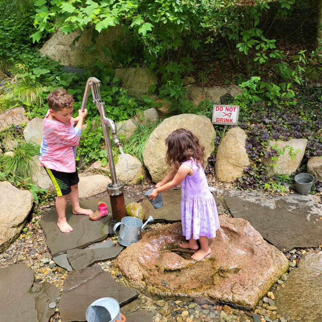 A 6-year-old kid with short brown hair in a salmon-colored tie-die shirt and black shorts operates a water pump. The water coming out of the pump is coming near a small galvanized metal bucket being held by a 3-year-old kid with long curly dark brown hair wearing a purple tie-dyed dress. Both kids are barefoot. They are standing on rocks and slate in a shallow water feature, with rocks and greenery in the background.
