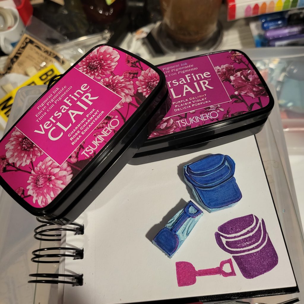 Two VersaFine Clair inkpads, in pink and purple, dominate the top half of the image. They rest on a small spiral notebook with unruled pages. On the lower half of the notebook page are 2 hand-carved, blue stamps depicting a beach shovel and bucket. In the lower right quadrant of the page the stamps have been impressed on the page, showing a pink shovel laying horizontally just to the left of a purple bucket.