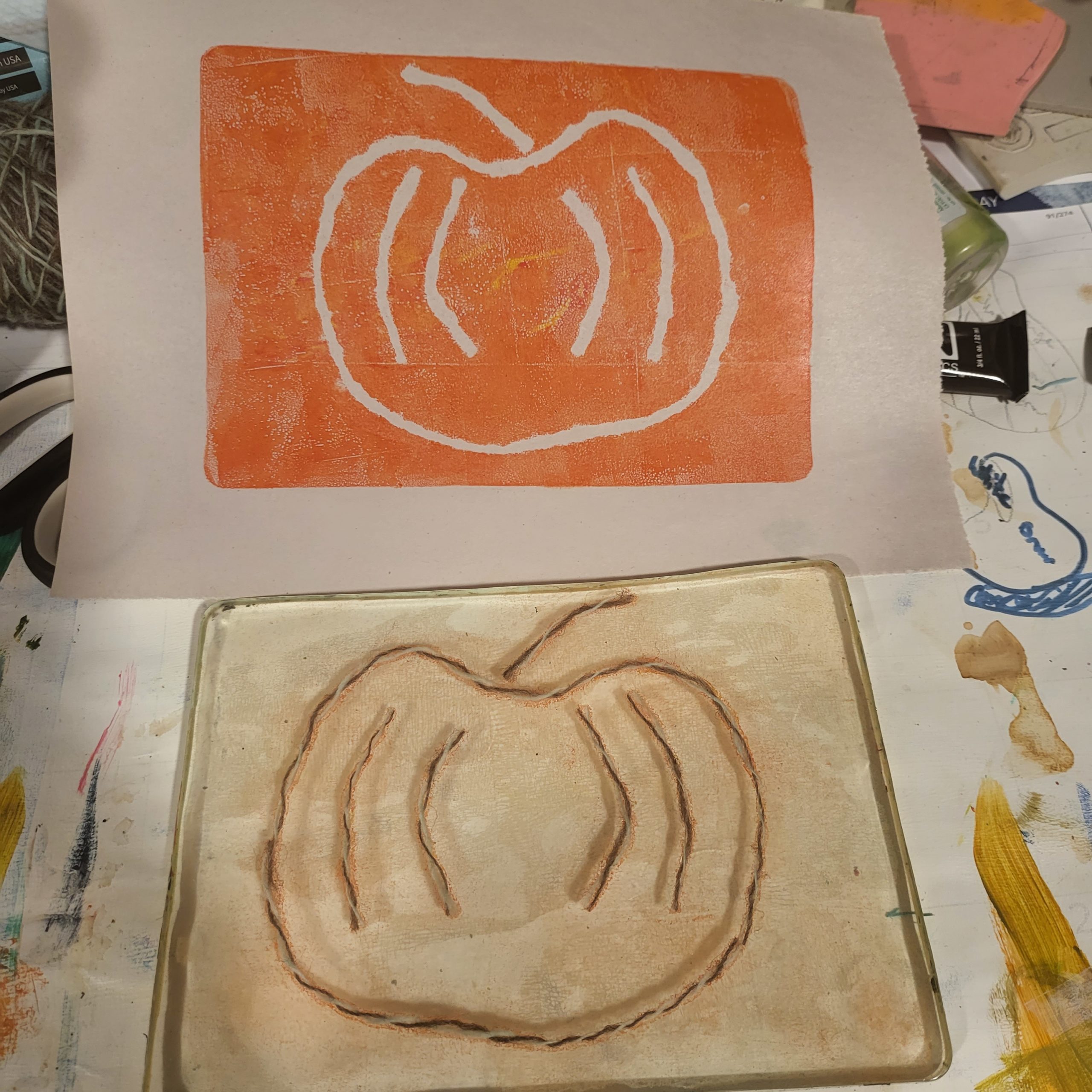 The bottom half of this image shows a gelatin plate with yarn on it, forming a simple pumpkin shape. The top half is the print that just came off the same gelatin plate - an orange field with a pumpkin shape outline that is the color of the underlying paper.