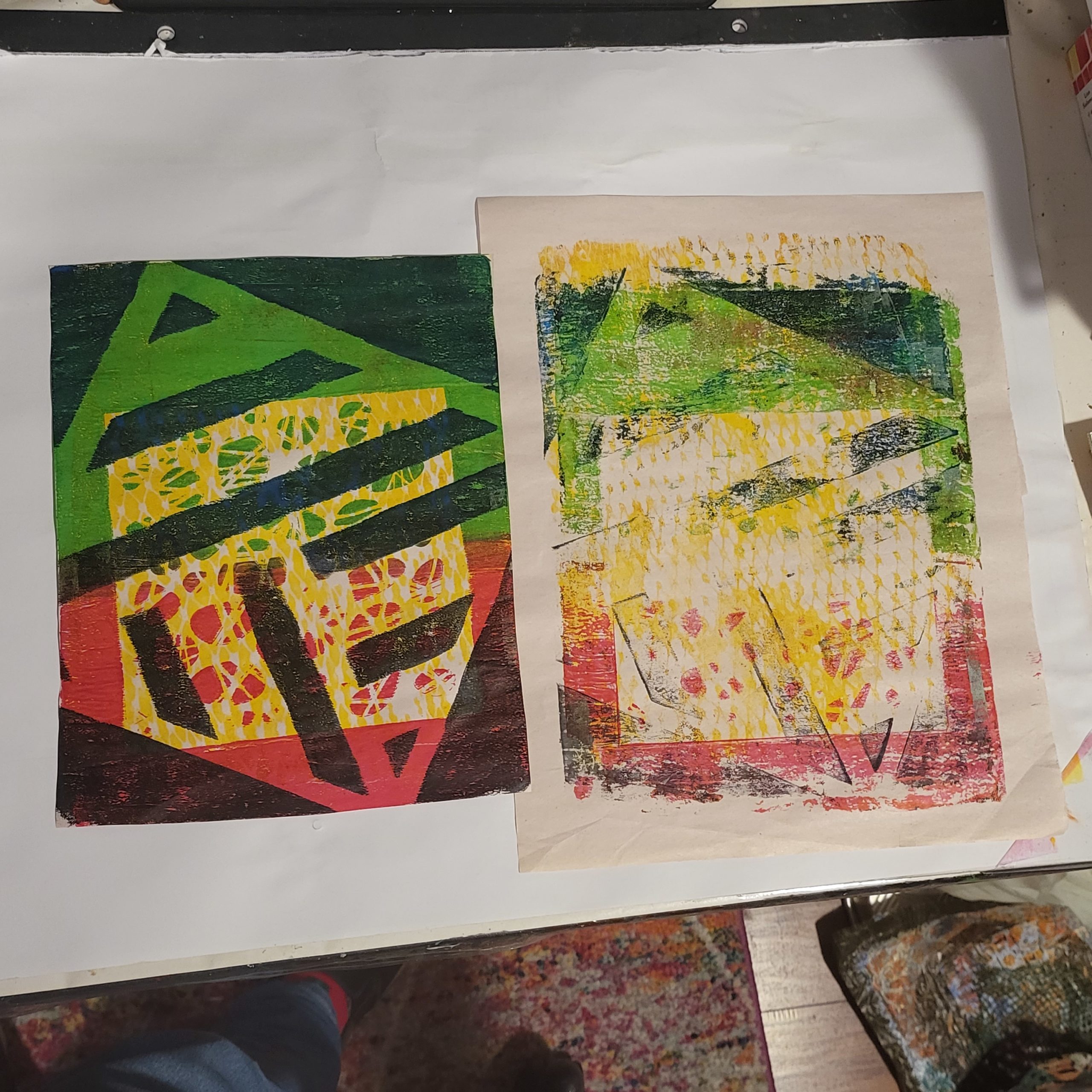 This image has 2 monoprints in it - the monoprint on the right is a mess of layers that did not work out - 2 stencils and netting, with no background so a lot of the original paper is showing through. The monoprint on the left has a yellow mask of a stencil, on a green and red background, with another stencil layered on top in black. Experiments help us determine what works and what does not, so that we can produce what we want to.