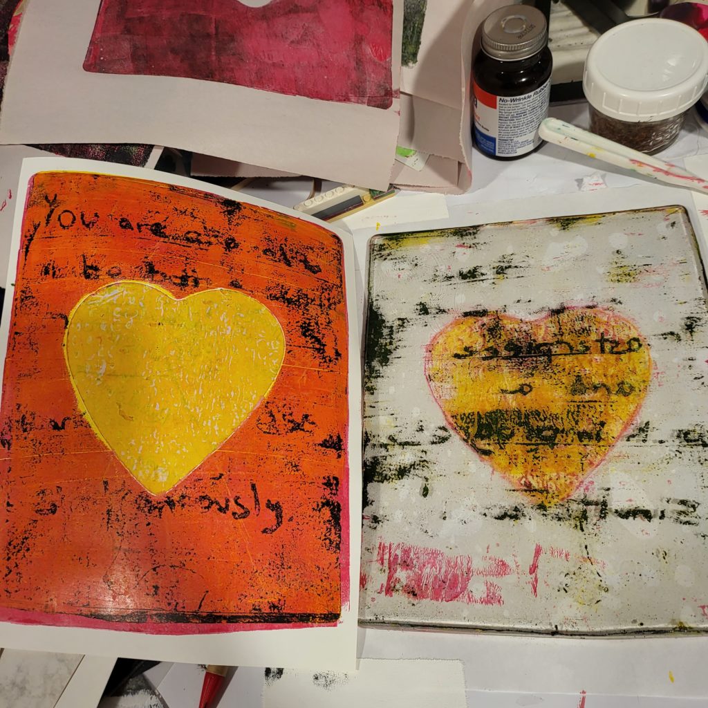 The right half of this image is a gelatin plate with a ghost image - some writing, especially in the middle where there is a yellow heart mask. The mask ended up splitting, and the top part of the heart-shaped mask ended up on the print. The print takes up the left half of the image, with a plain yellow heart in the middle, on an orange background, with some black text that did not come off the gelatin plate in a readable manner. This is a failed experiment.